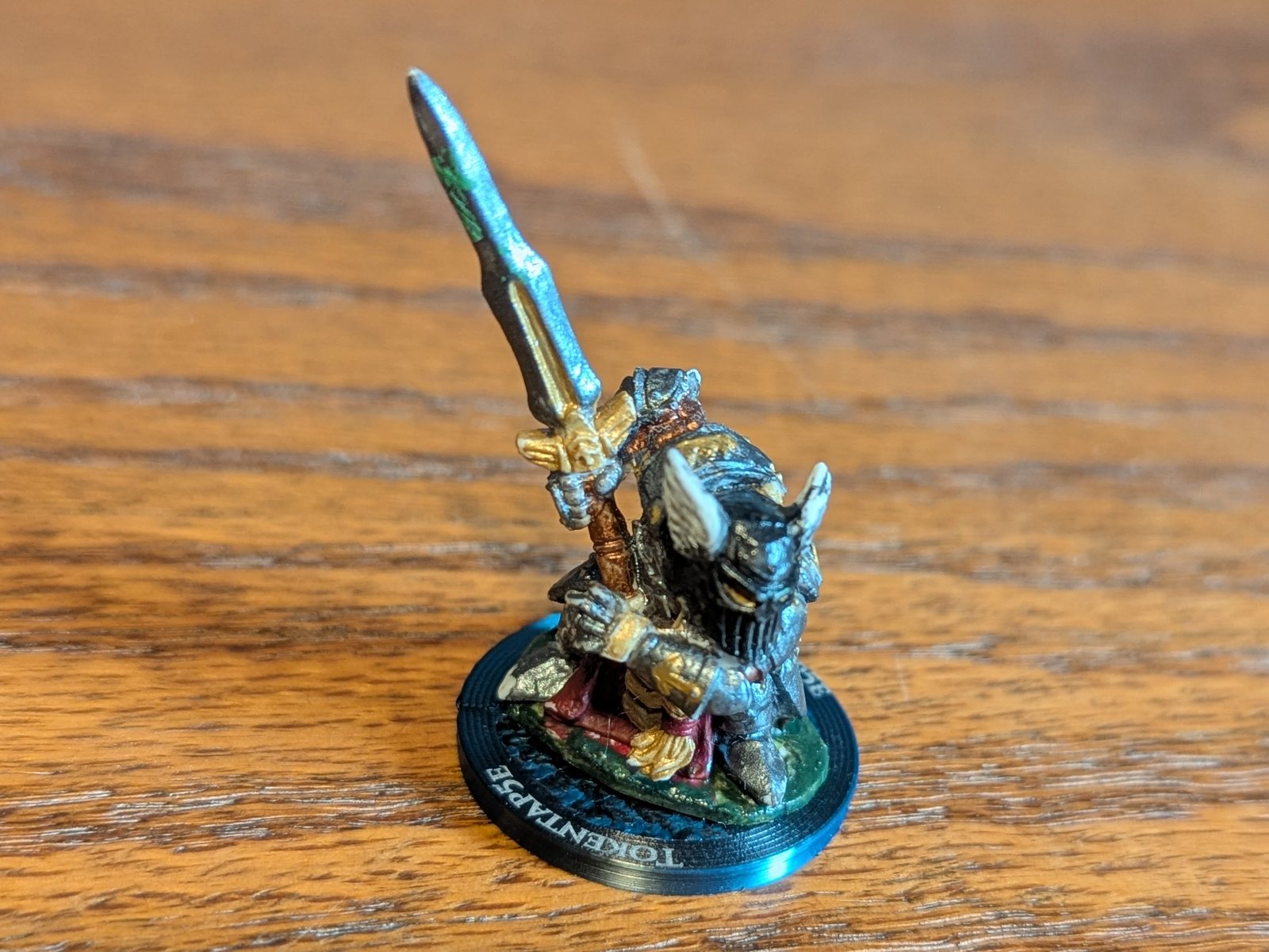 A painted miniature of an armored warrior with a raised sword, standing on a TokenTap5e coin on a wooden table, the name engraved around the coin's rim.