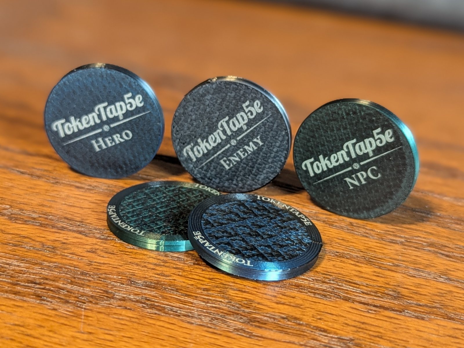Five TokenTap5e miniature coins on a wooden table. Three stand upright showing their laser-engraved faces: the TokenTap5e wordmark over a d20 divider, marked Hero, Enemy and NPC. Two lie flat showing the back, with TokenTap5e engraved twice around the rim.