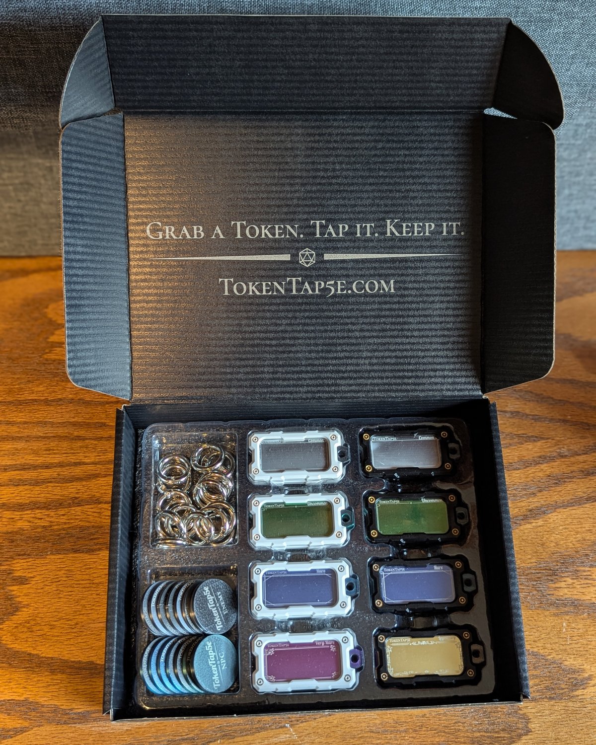 The Core Pack open and seen straight on, sixteen NFC item tokens in black and white housings, hang rings and two stacks of engraved coins in the tray, the lid engraved with Grab a Token. Tap it. Keep it.