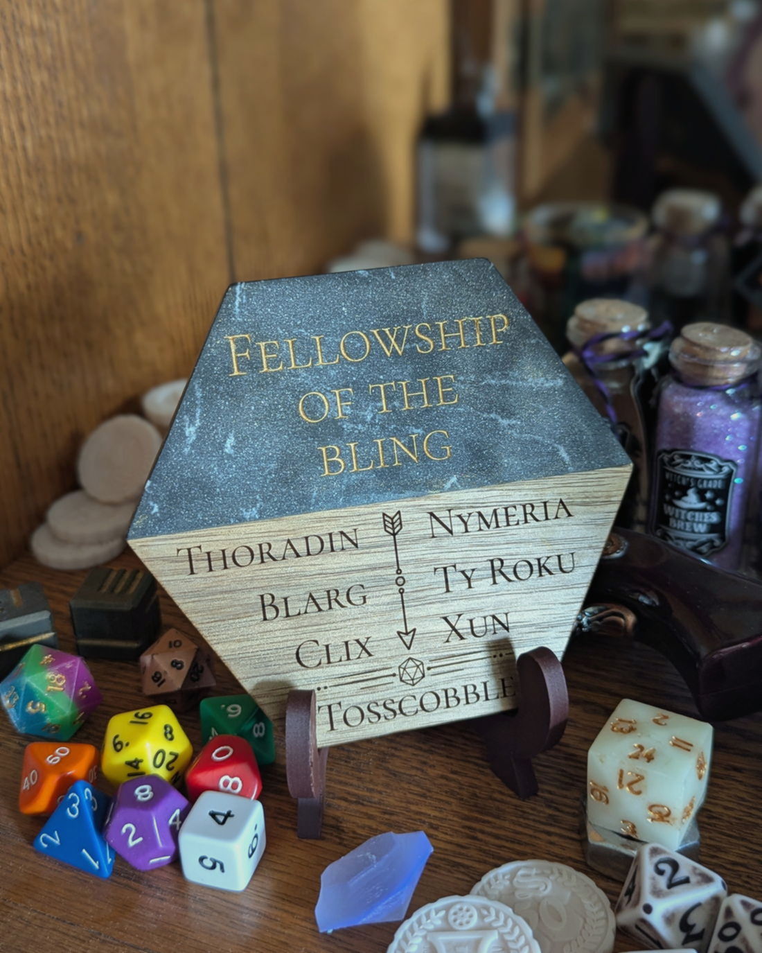 A hexagonal Campaign Crest on an easel, standing on a shelf among dice, miniatures and potion bottles. The upper half is dark marbled resin engraved in gold with the party name Fellowship of the Bling, the lower half is engraved wood listing seven character names.