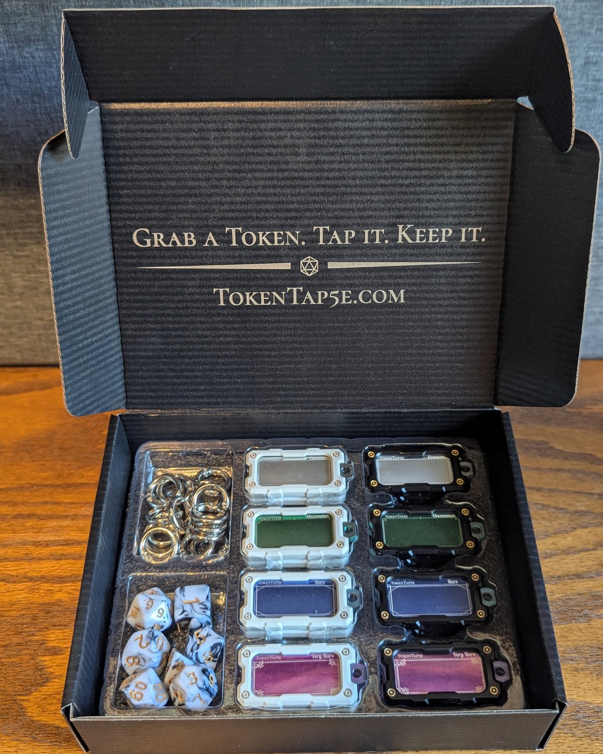 The Expansion Pack open and seen straight on, sixteen NFC item tokens in black and white housings, hang rings and a black and white marbled dice set with gold numbers in the tray, the lid engraved with Grab a Token. Tap it. Keep it.