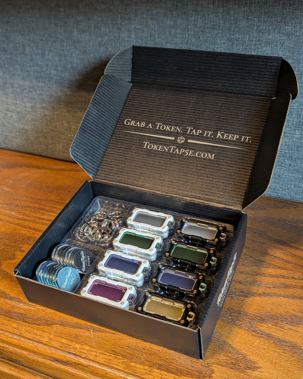 An open TokenTap5e Core Pack on a table, sixteen NFC item tokens, hang rings and engraved hero, NPC and enemy coins seated in a molded tray, the lid engraved with Grab a Token. Tap it. Keep it.